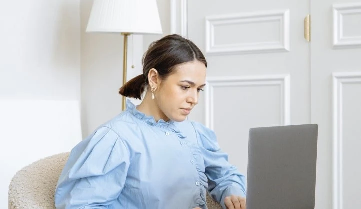 Mujer con cabello castaño en cola de caballo usando una laptop en una habitación blanca con una lámpara dorada